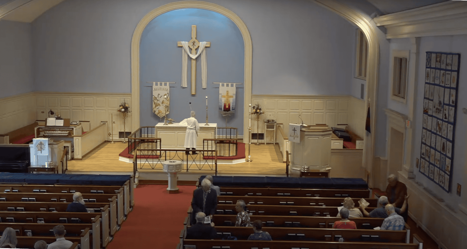 A church interior with a person in robes near the altar. Several people are seated in pews, and a cross hangs on the wall.